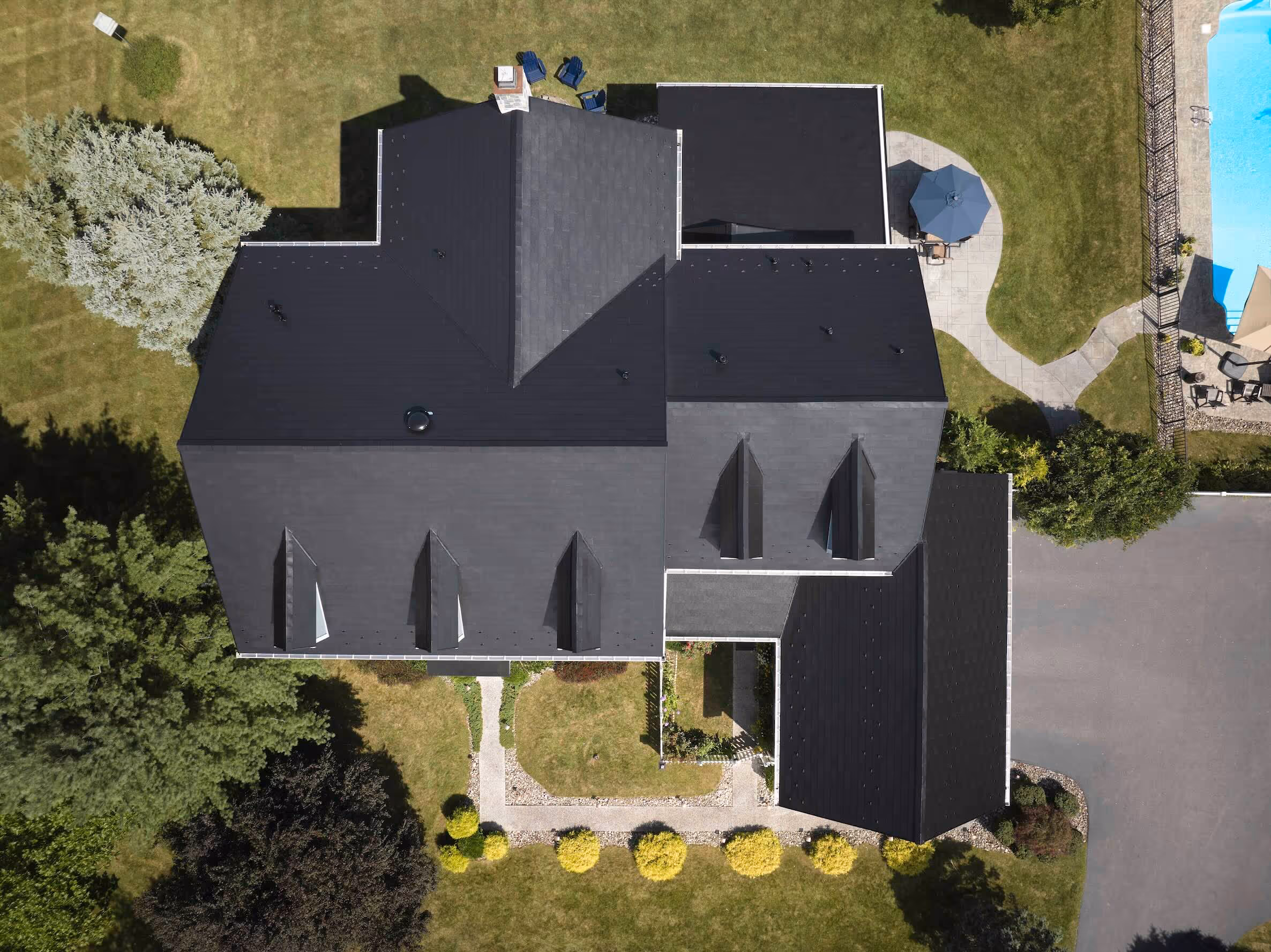 Large two-story suburban house with blue siding, black shutters, stone accents, and a dark metal Timbersteel roof surrounded by green landscaping.